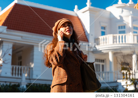 Mid shot of a elegant Caucasian woman in a coat and hat calling by the smartphone and smiling. Beautiful city street in background. Bottom view. Modern communication 131304161