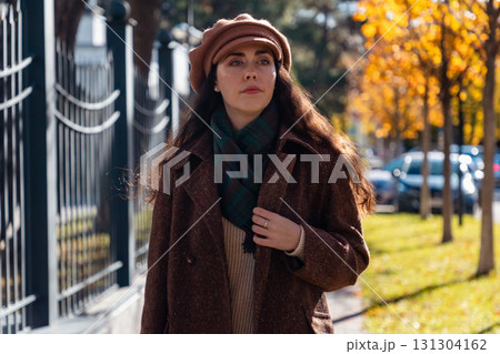 Mid shot of pensive young Caucasian woman in a hat and coat walking along an autumn street in a sunny city. Concept of psychology Mid shot of pensive young Caucasian woman in a hat and coat walking along an autumn street in a sunny city. Concept of psychology 131304162