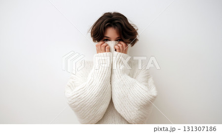 Young woman in oversized white sweater playfully hiding her face while looking at the camera against a plain light background 131307186