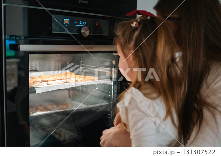 A child and caregiver enjoy watching cookies bake together in the warm oven, sharing a special moment A child and caregiver enjoy watching cookies bake together in the warm oven, sharing a special moment 131307522