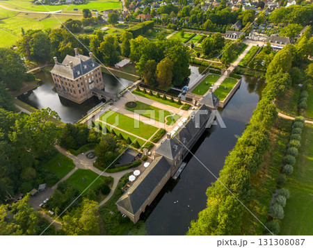 Aerial view of historic moated castle with gardens, bridge, and adjacent long building, surrounded by trees, water features, and a nearby village under warm sunlight. Aerial view of historic moated castle with gardens, bridge, and adjacent long building, surrounded by trees, water features, and a nearby village under warm sunlight. 131308087