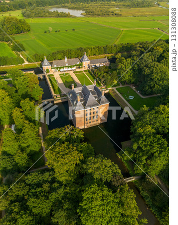 Aerial view of historic moated castle with gardens, bridge, and adjacent long building, surrounded by trees, water features, and a nearby village under warm sunlight. 131308088