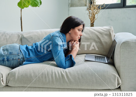 Young relaxed woman lying on sofa at home, looking at laptop 131311025