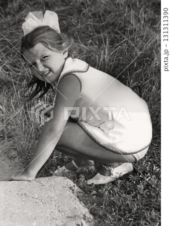 A vintage portrait of a seven-year-old girl in a short dress with a white bow in her hair, crouched and searching for something in the grass. Photo from 1974. A vintage portrait of a seven-year-old girl in a short dress with a white bow in her hair, crouched and searching for something in the grass. Photo from 1974. 131311390