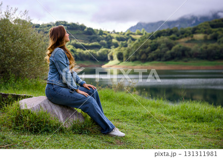 Young woman with braid sitting on rock near mountain lake 131311981