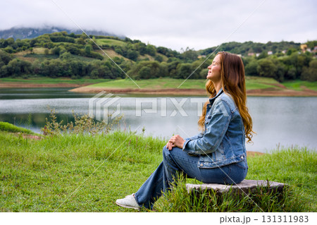 Young woman sitting by lake in denim jacket and jeans 131311983