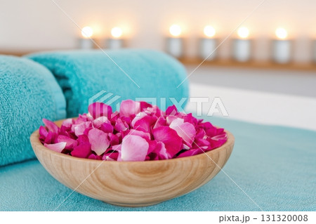 The calming wellness spa environment showcases plush turquoise towels, a wooden bowl filled with vibrant magenta flower petals, and softly glowing light blue candles, perfect for relaxation. 131320008