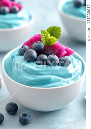 A colorful smoothie bowl features smooth turquoise spirulina, topped with magenta dragon fruit, fresh blueberries, and a vibrant mint leaf, presented on a light blue tabletop. 131320016