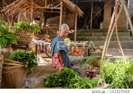 An elderly woman is at the market, surrounded by fresh produce and colorful items 131325584