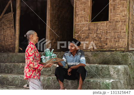 An elderly woman serves tea to a young man in a traditional Indonesian village An elderly woman serves tea to a young man in a traditional Indonesian village 131325647