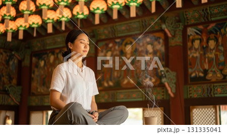 Asian woman meditating with closed eyes and burning incense in traditional Korean Buddhist temple with colorful dancheong paintings and lotus lanterns Asian woman meditating with closed eyes and burning incense in traditional Korean Buddhist temple with colorful dancheong paintings and lotus lanterns 131335041