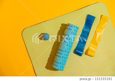 Flat lay on yoga mat with foam roller blue and yellow bands and massage ball over yellow backdrop active recovery 131335191