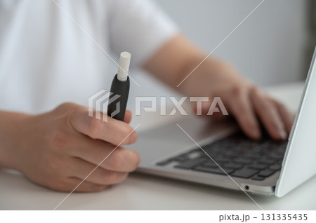 A woman smokes an electronic cigarette while working on a laptop. Tobacco heating system. A woman smokes an electronic cigarette while working on a laptop. Tobacco heating system. 131335435