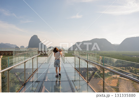 Caucasian woman holds her little son in her arms while standing on a glass bridge in the mountains.  131335880