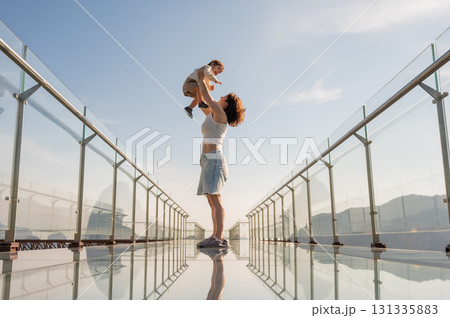 Caucasian woman holds her little son in her arms while standing on a glass bridge in the mountains. Caucasian woman holds her little son in her arms while standing on a glass bridge in the mountains. 131335883