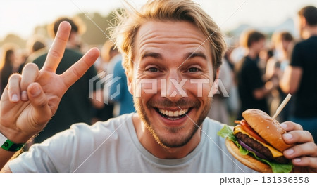 Happy young man eating a burger and showing a peace sign at a summer festival. Portrait of a smiling person enjoying fast food outdoors at sunset 131336358