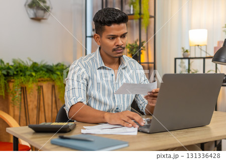 Indian man uses laptop and calculator to enter bill numbers compare sums plan budget savings finance 131336428