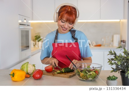 Young woman with headphones plays anti stress music cuts cucumbers and tomatoes for fresh salad prep 131336434