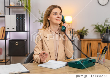 Businesswoman calls client on retro phone spins rotary dial takes notes agreement formal tone smile Businesswoman calls client on retro phone spins rotary dial takes notes agreement formal tone smile 131336458