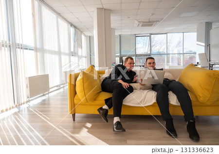Two Caucasian men discussing work on a laptop while sitting on a sofa. Modern office.  131336623