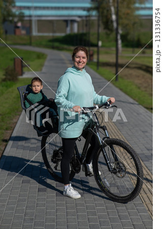 Caucasian woman riding bicycle with toddler son sitting behind her in child seat in park. Vertical photo. Caucasian woman riding bicycle with toddler son sitting behind her in child seat in park. Vertical photo. 131336754