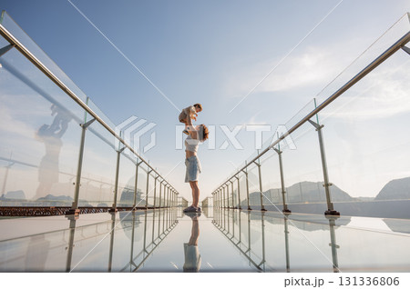 Caucasian woman holds her little son in her arms while standing on a glass bridge in the mountains. Caucasian woman holds her little son in her arms while standing on a glass bridge in the mountains. 131336806