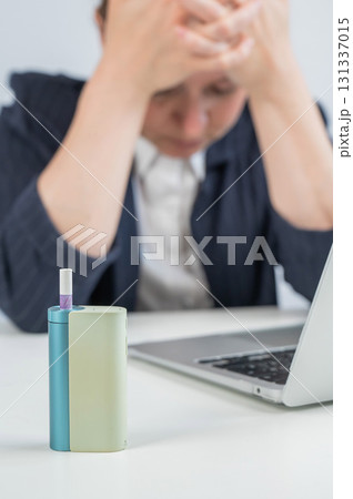 Tired businesswoman smoking an electronic cigarette while working on a laptop. Tobacco heating system. 131337015