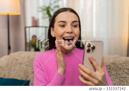 Young woman on sofa looking at mobile phone screen opening mouth shocked by unexpected surprise 131337142