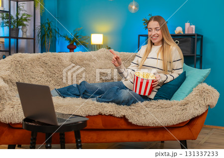 Caucasian woman enjoying movie on sofa eating popcorn laughing watching laptop indoors smiling happy Caucasian woman enjoying movie on sofa eating popcorn laughing watching laptop indoors smiling happy 131337233
