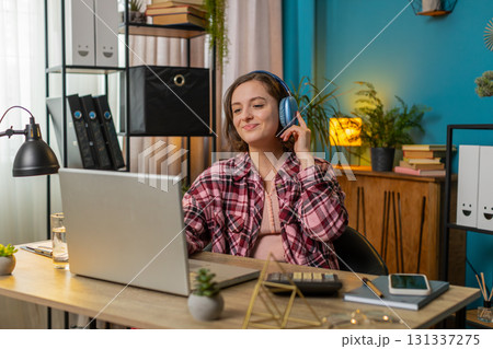 Excited business woman listening music through wireless headphones sitting at home office table Excited business woman listening music through wireless headphones sitting at home office table 131337275