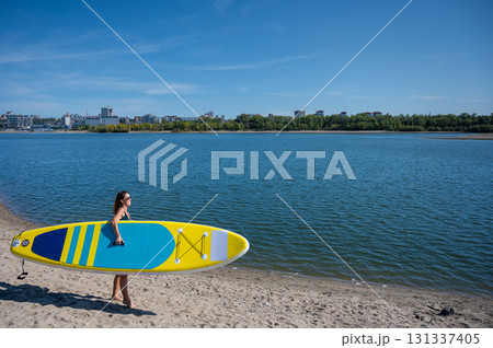 Caucasian woman walks along the beach and carries a sup board on the river in the city. Summer sport. 131337405