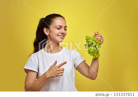 portrait of attractive Caucasian smiling woman holds grapes isolated on yellow studio background 131337811