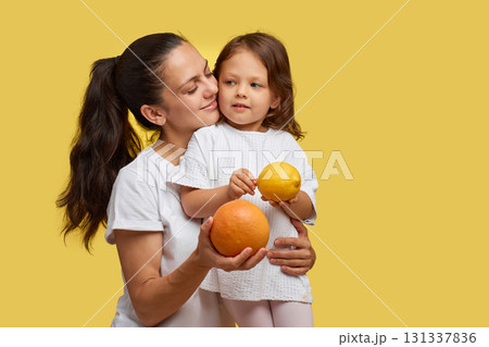 Cheerful little child girl with mother holding lemon and grapefruit on yellow background. happy family having fun together Cheerful little child girl with mother holding lemon and grapefruit on yellow background. happy family having fun together 131337836