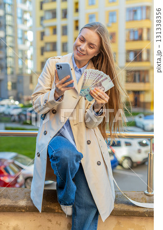 Happy rich mature businesswoman lady using smartphone, holding money hryvnias cash on city street Happy rich mature businesswoman lady using smartphone, holding money hryvnias cash on city street 131338356