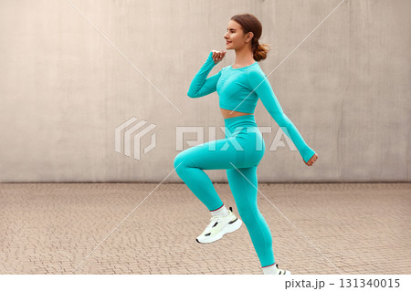 Fitness. Jumping Woman Doing Workout At Outdoor Stadium. Side View Of Fit Girl In Fashion Sporty Outfit Doing Exercise Against Concrete Wall. Dynamic Movement. 131340015