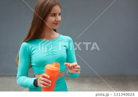 Fit woman dressed in turquoise activewear, holding an orange shaker bottle, smiling during outdoor exercise, embodying health and vitality. Workout concept Fit woman dressed in turquoise activewear, holding an orange shaker bottle, smiling during outdoor exercise, embodying health and vitality. Workout concept 131340028