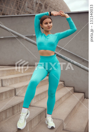 Fit woman in turquoise activewear poses confidently on outdoor stairs, showcasing strength and vitality in a dynamic fitness environment. Workout concept 131340033