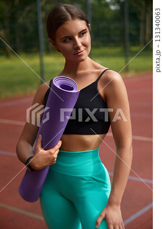 Fit young woman with a purple yoga mat stands confidently on a sports court, dressed in a black crop top and teal leggings, embodying strength and wellness in a vibrant workout environment. Fit young woman with a purple yoga mat stands confidently on a sports court, dressed in a black crop top and teal leggings, embodying strength and wellness in a vibrant workout environment. 131340063