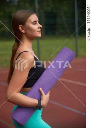 Young woman with long hair holds a purple yoga mat while standing on a sports court, dressed in athletic wear, embodying fitness and wellness in an outdoor workout environment. Young woman with long hair holds a purple yoga mat while standing on a sports court, dressed in athletic wear, embodying fitness and wellness in an outdoor workout environment. 131340064