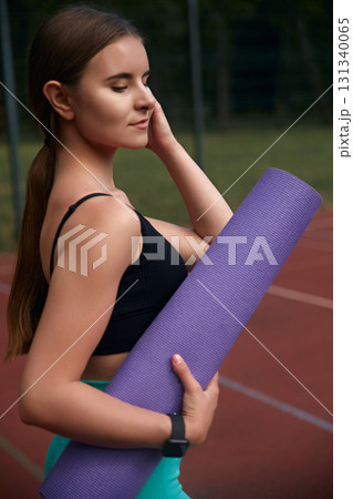 Fit young woman with long hair holds a purple yoga mat while standing on a sports field, showcasing a healthy lifestyle and dedication to fitness and wellness in an outdoor environment. Fit young woman with long hair holds a purple yoga mat while standing on a sports field, showcasing a healthy lifestyle and dedication to fitness and wellness in an outdoor environment. 131340065