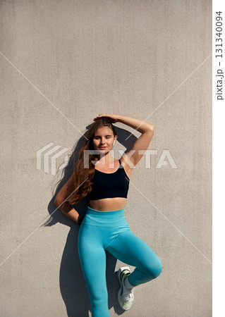 Fit woman in athletic wear poses confidently against a textured wall, showcasing strength and determination, embodying the spirit of fitness and wellness in a vibrant workout environment. 131340094