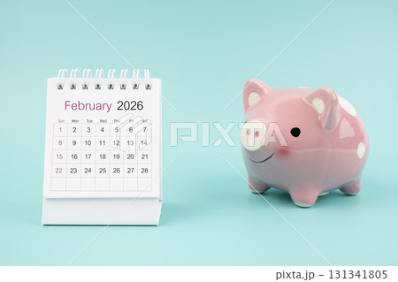 February 2026 Desk calendar and pink piggy bank on blue background. February 2026 Desk calendar and pink piggy bank on blue background. 131341805