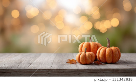 Three pumpkins of varying sizes sit on a weathered wooden table alongside fallen autumn leaves, creating a warm and inviting autumn scene with soft bokeh lights in the background 131342939