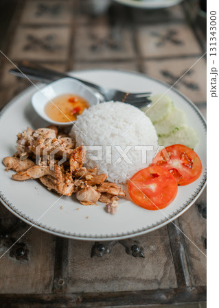 A plate of Thai chicken rice with fresh tomatoes, cucumbers, and spicy dipping sauce, served on a rustic wooden table. A plate of Thai chicken rice with fresh tomatoes, cucumbers, and spicy dipping sauce, served on a rustic wooden table. 131343000