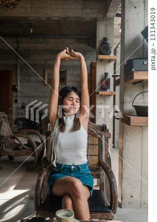 A young woman stretches in a sunlit industrial-style loft, surrounded by vintage decor and rustic furniture. 131343019
