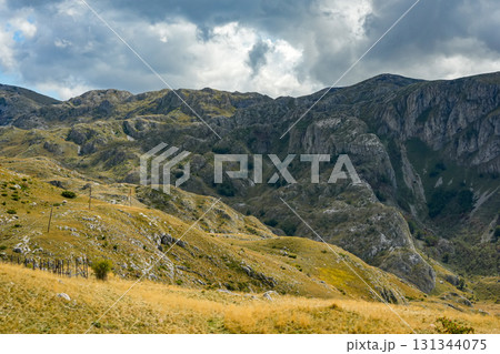 Scenic road winding through golden hills and mountains with a car visible in the distance under a bright blue sky with white clouds. Ideal for travel, landscape, adventure, and nature projects 131344075
