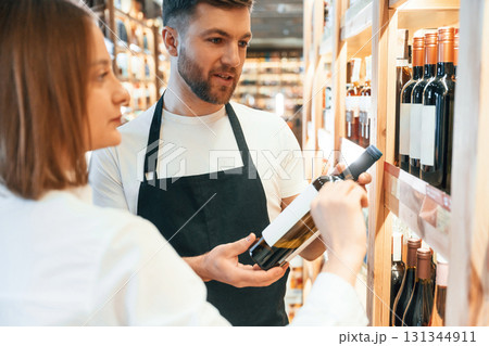 Asking for help. The shop owner helps a young woman to choose a good wine Asking for help. The shop owner helps a young woman to choose a good wine 131344911