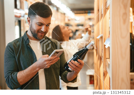 Smartphone and bottle in hands of a guy. Man and woman are choosing wine in the shop 131345312