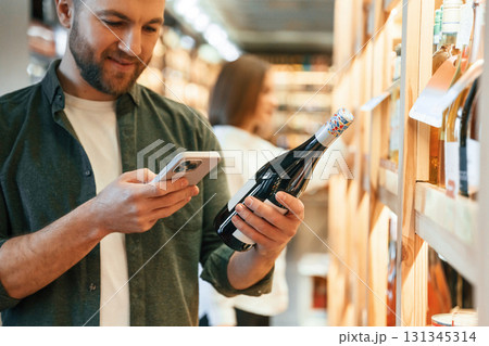 Smartphone and bottle in hands of a guy. Man and woman are choosing wine in the shop Smartphone and bottle in hands of a guy. Man and woman are choosing wine in the shop 131345314