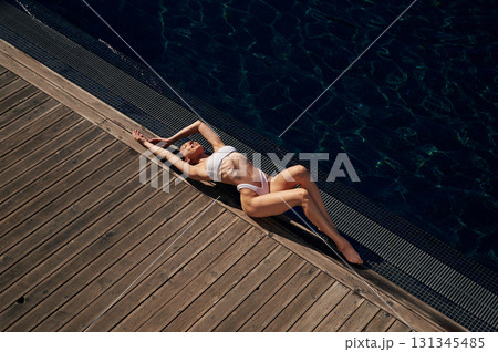 Lying down, enjoying sunlight. Woman in white bikini is near swimming pool 131345485